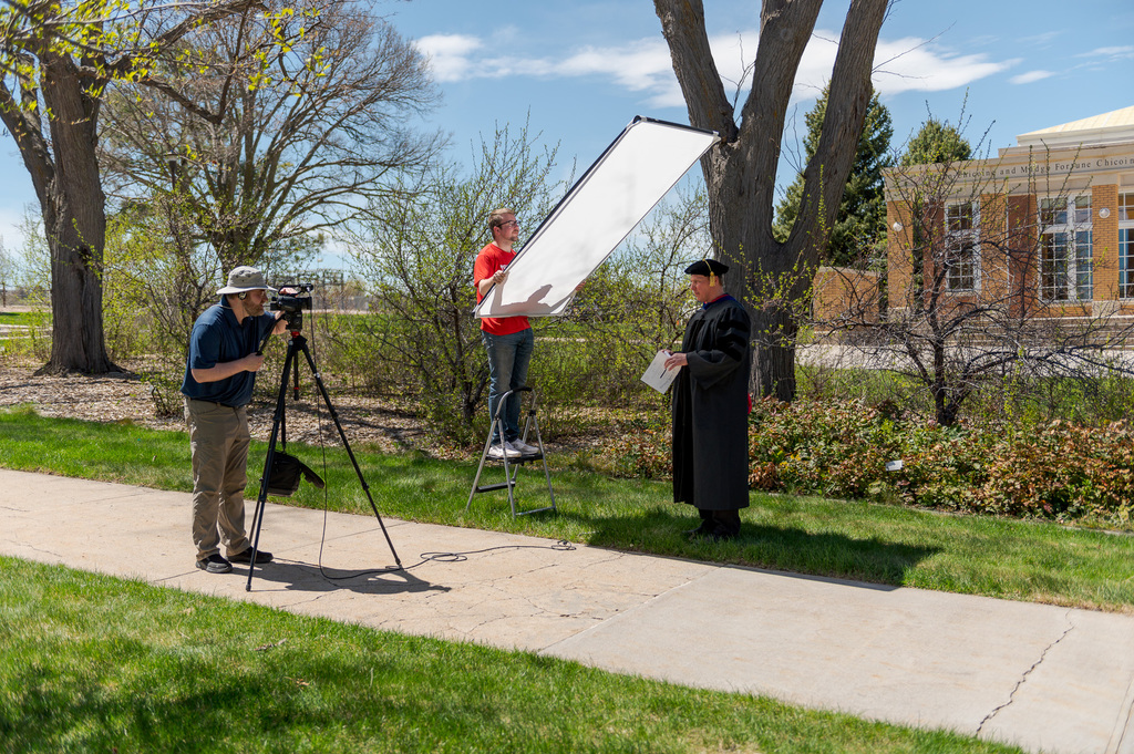 Dr. Kurt Kinbacher, associate professor prepares to deliver his message to the graduating class while Daniel Binkard adjusts the camera