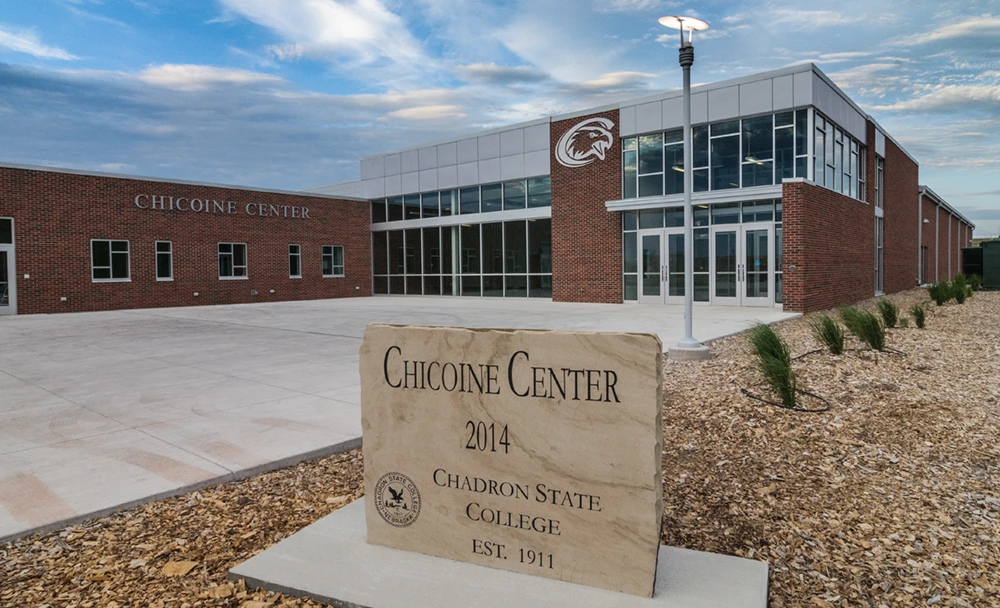 An athletic building of brick, metal, and glass, with a sign in the foreground reading Chicoine Center, 2014, Chadron State College, Est. 1911