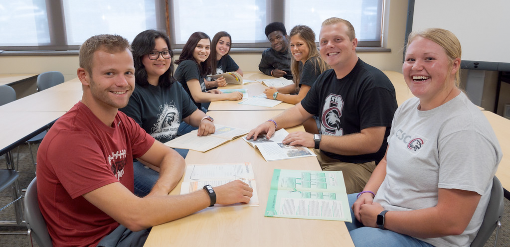 A group of students sitting all around a table smiling.